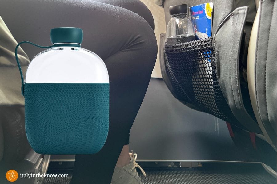 Knee level view of a seated person on a plane with a water bottle in the seat back storage compartment and a water bottle overlay