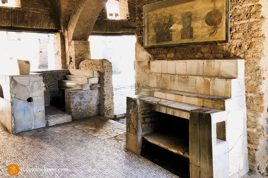 Popina interior with mosaic floor, sales counter, shelves for displaying food, and a painting on the wall of foods available for purchase